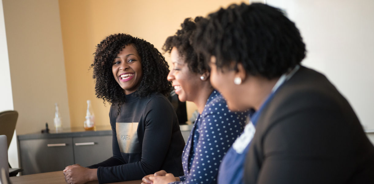 A group of 3 diverse women sat at a board table, one of whom is laughing