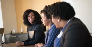 A group of 3 diverse women sat at a board table, one of whom is laughing